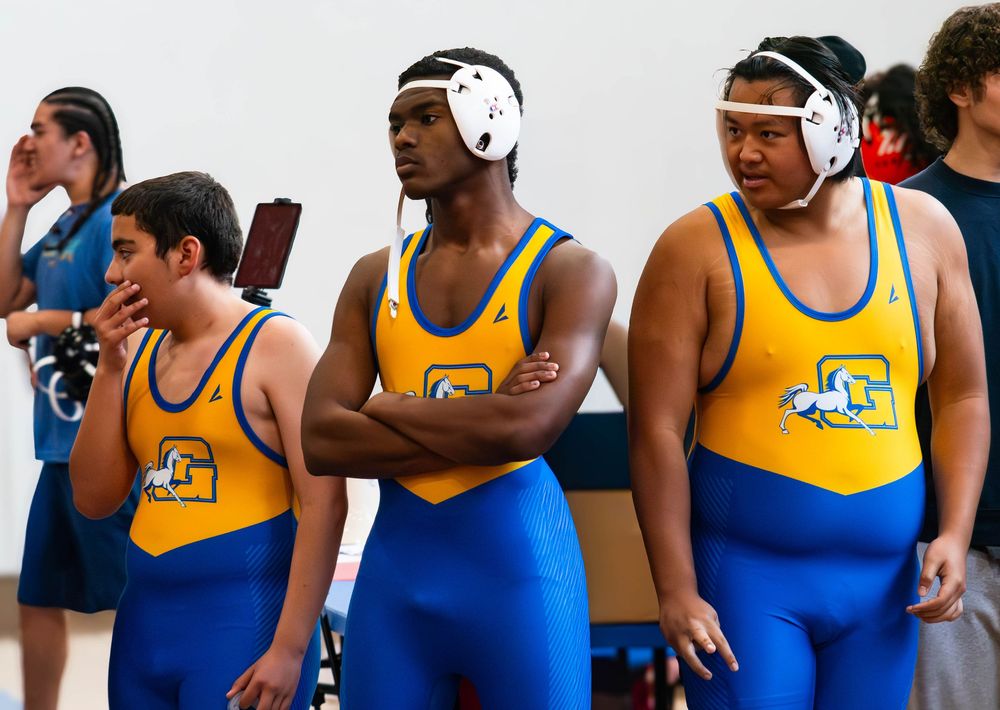 High school wrestlers in blue and gold uniforms stand together wearing headgear during a match.