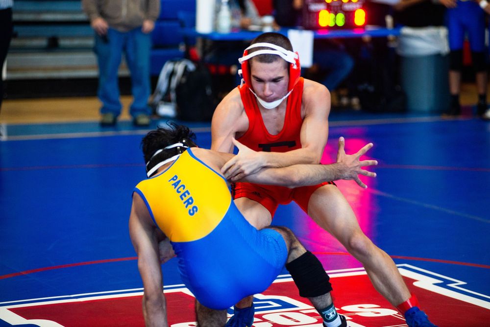 Wrestlers in red and blue uniforms grappling during a competitive match on a blue wrestling mat.