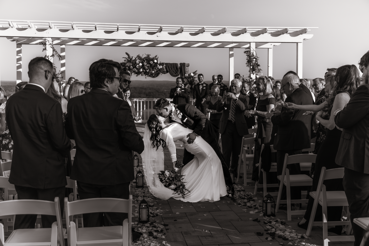 A wedding ceremony taking place outdoors under a white pergola with gathered guests.