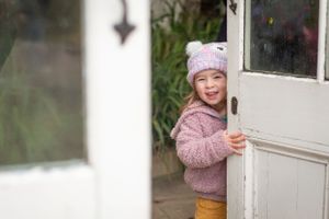 Child in pink sweater and hat peeking through white doorway with joyful expression.