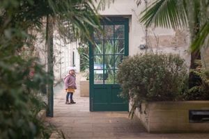Small figure walking through greenhouse corridor with green door and tropical plants.
