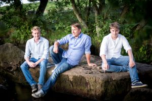 Three people sitting on large stone wall in wooded area wearing casual clothing.