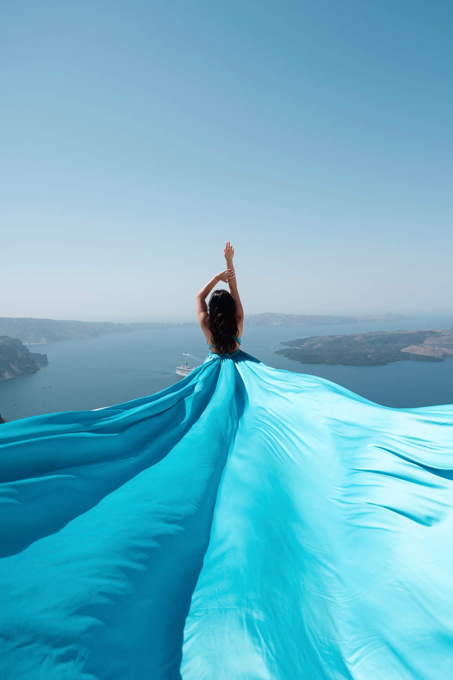 Flowing turquoise fabric creates dramatic silhouette against Mediterranean coastal landscape.