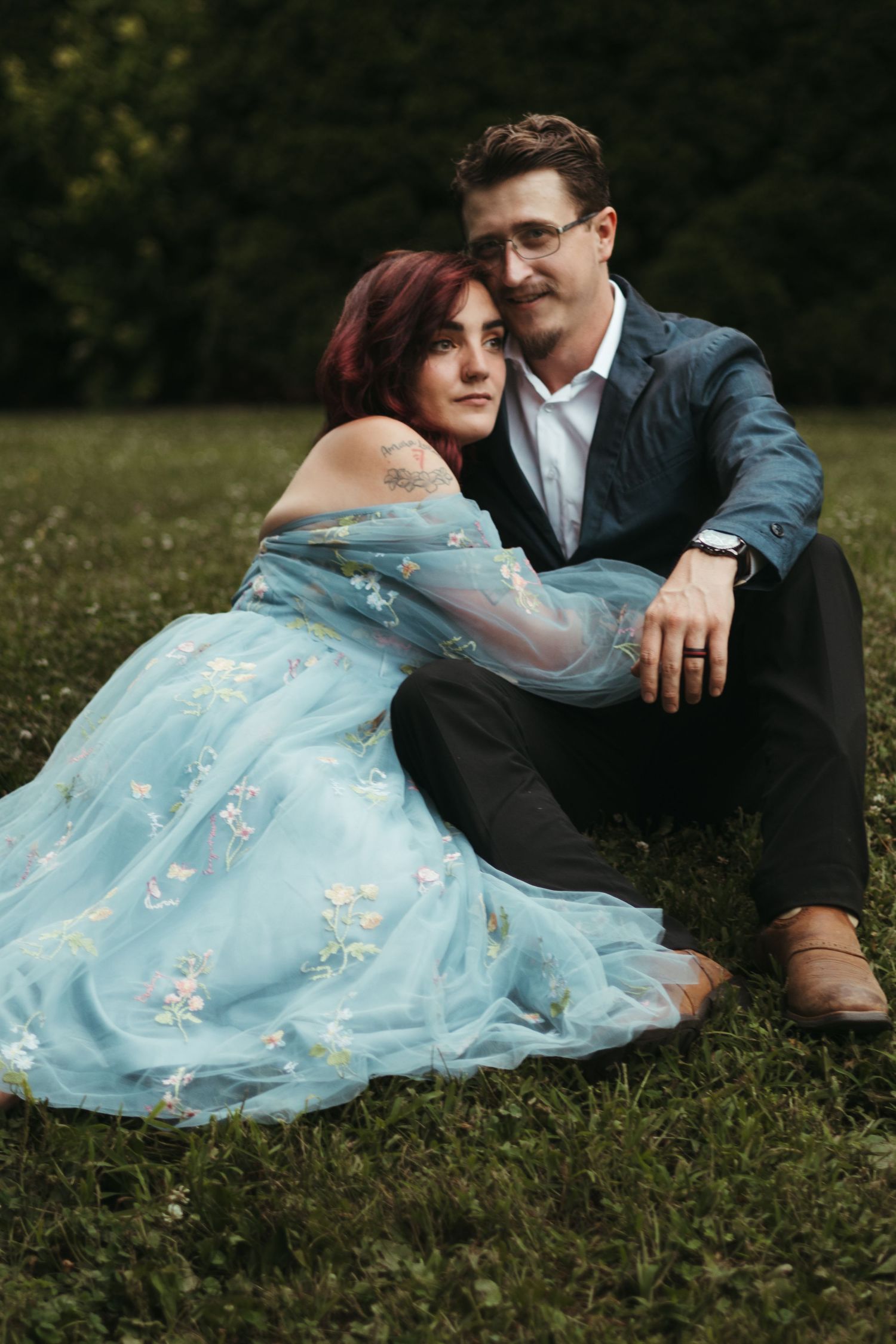 a couple sits in the grass smiling softly and looking into the distance. She's wearing a blue fairytale like dress