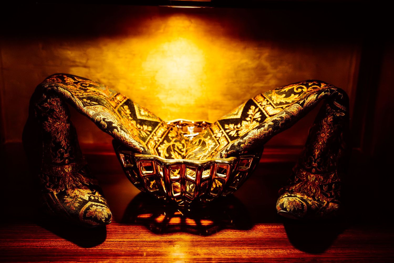 Golden ornate decorative bowl with intricate lattice design, flanked by two elaborate handles, illuminated by warm amber light.
