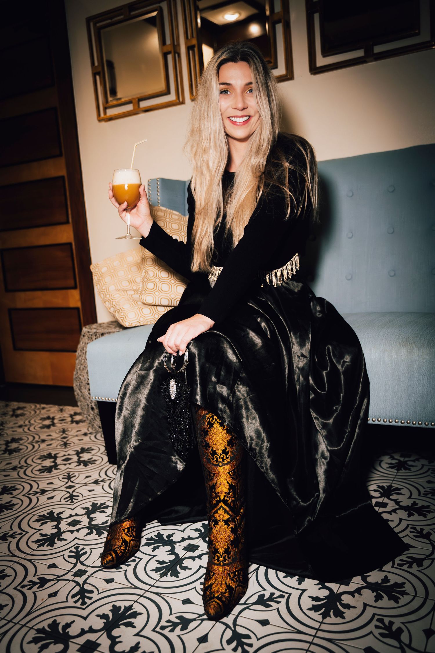 Elegant woman in black gown holding a beer glass, seated on a sofa with patterned tile floor.