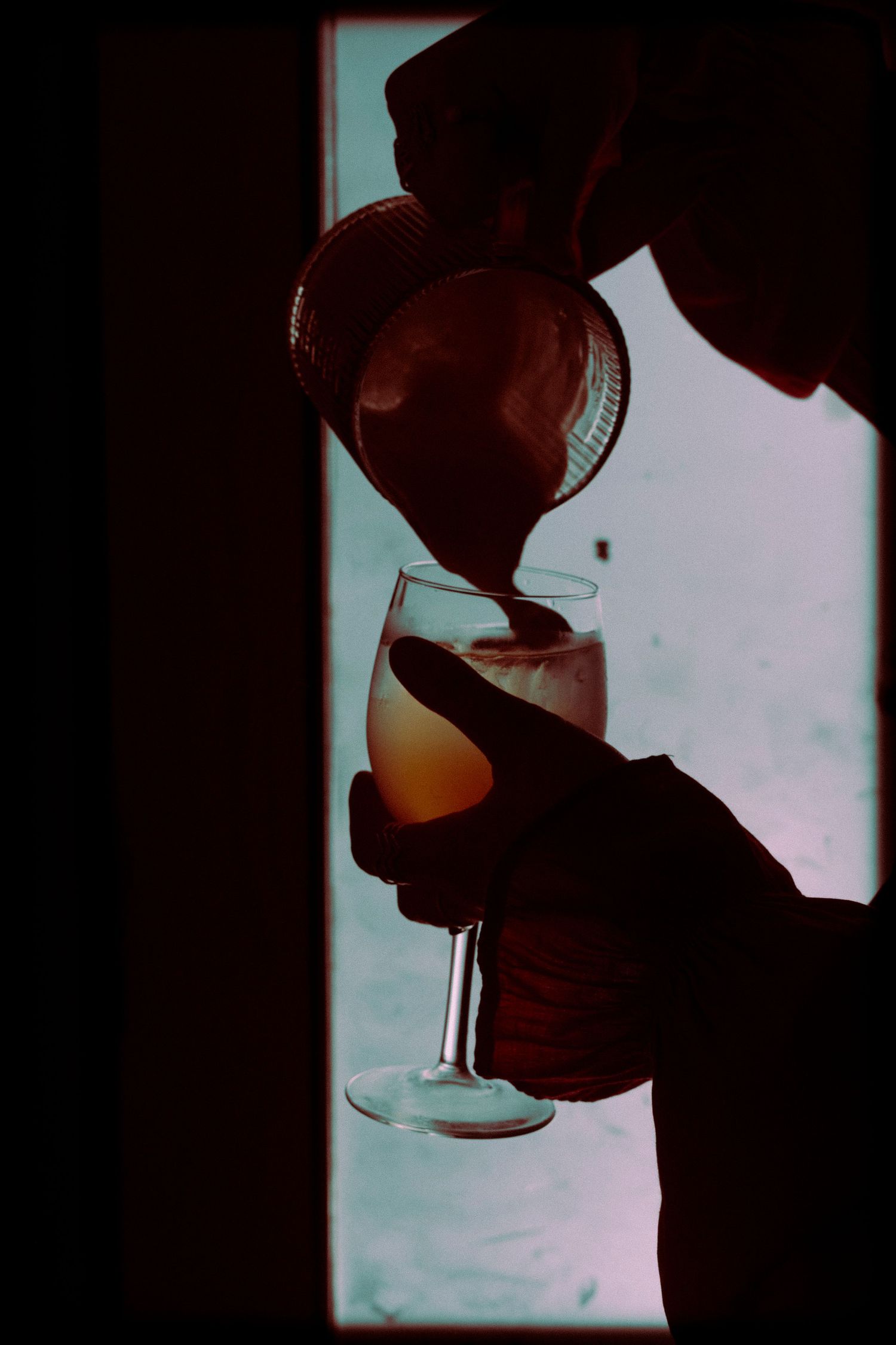 Silhouette of hands pouring a cocktail into a wine glass backlit by a window in moody lighting.