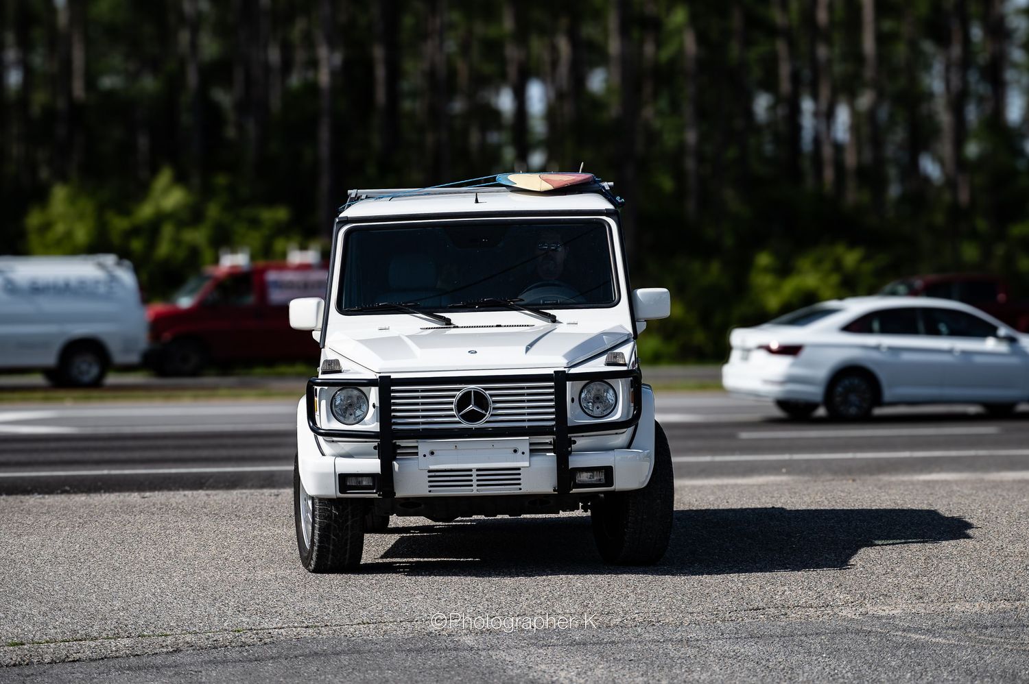 White Mercedes G-Wagon luxury SUV parked in a lot with cars and trees in background.