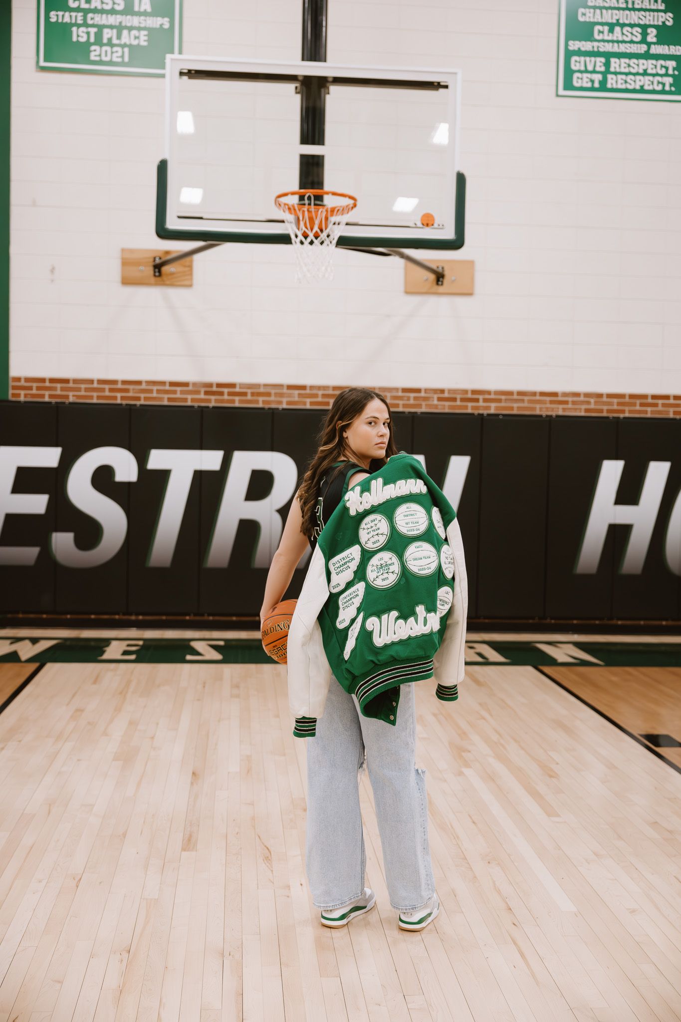 Young woman in green letterman jacket holding basketball on hardwood court in school gymnasium.