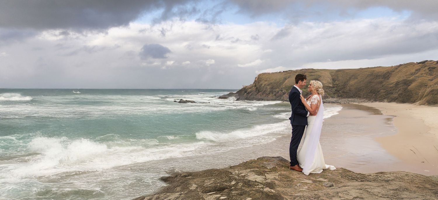 Bride and groom embrace romantically on coastal rocks with dramatic stormy ocean waves behind them.