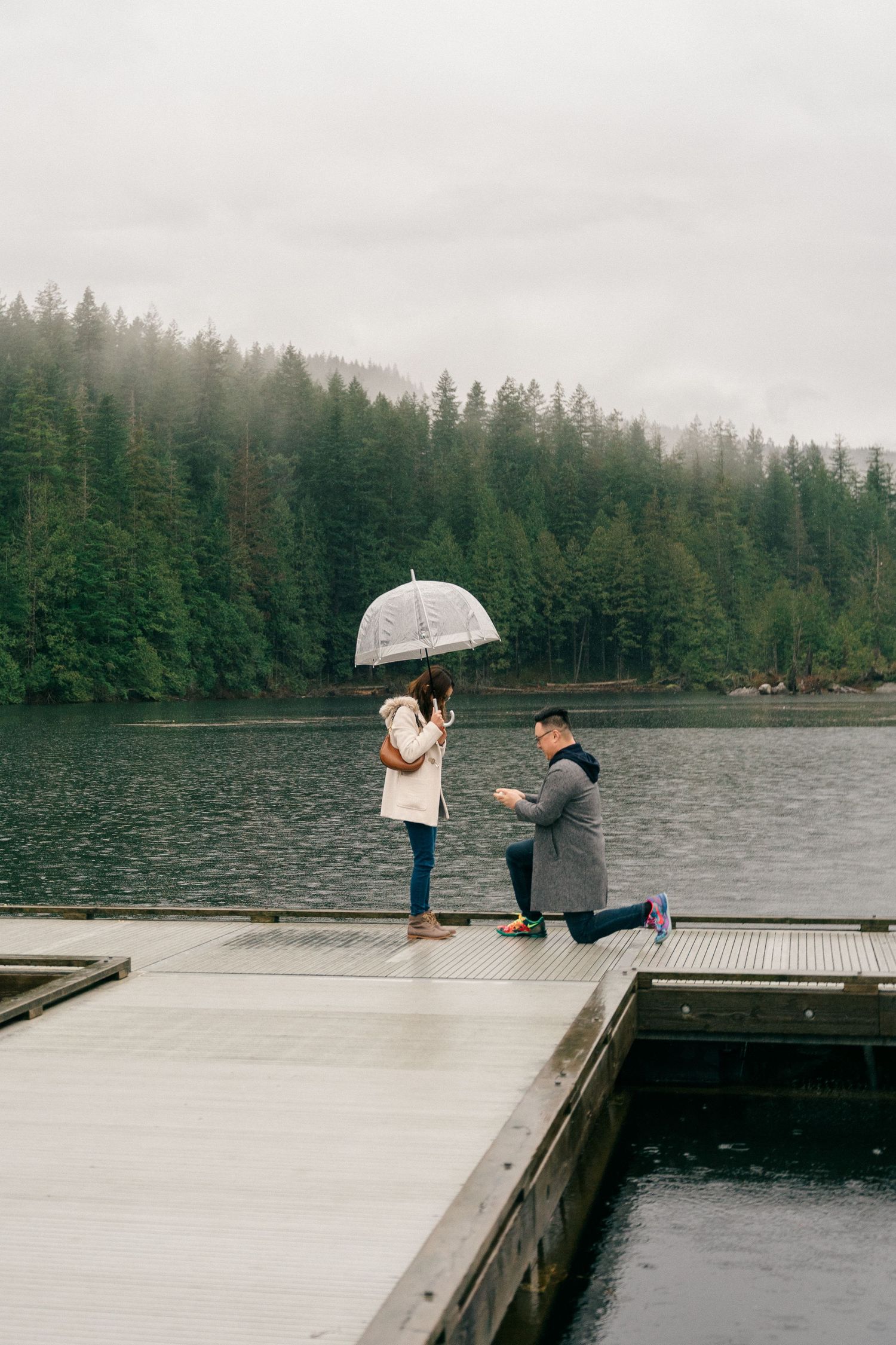 A romantic proposal moment unfolds on a misty wooden dock by a lake surrounded by evergreen forest.