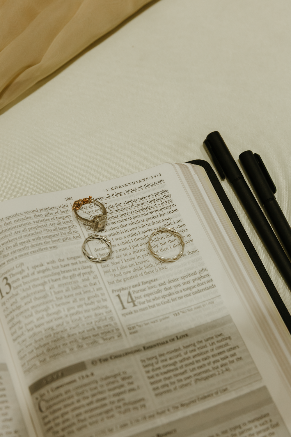 Three wedding rings resting on open Bible at 1 Corinthians passage with pens nearby.