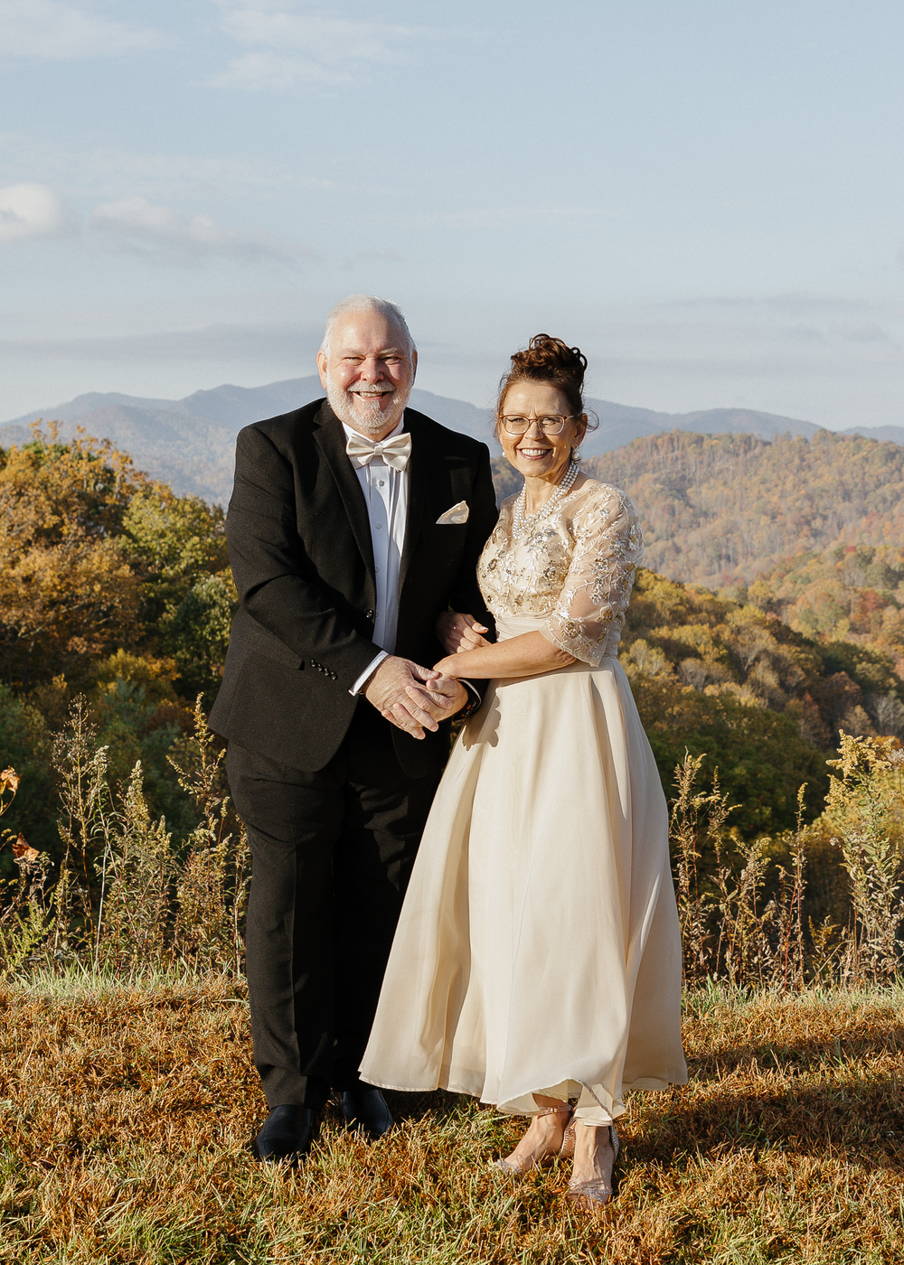Smiling bride and groom posing together in formal wedding attire with mountain views.
