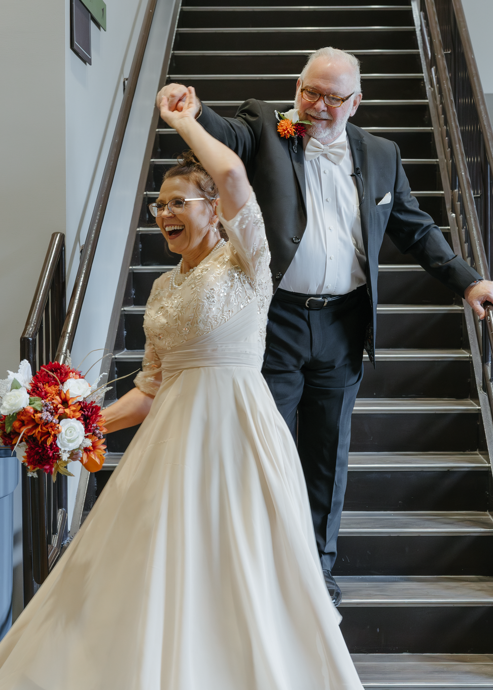 Joyful bride and groom celebrating on staircase, bride in white gown raising fist, groom in black tuxedo with boutonniere.