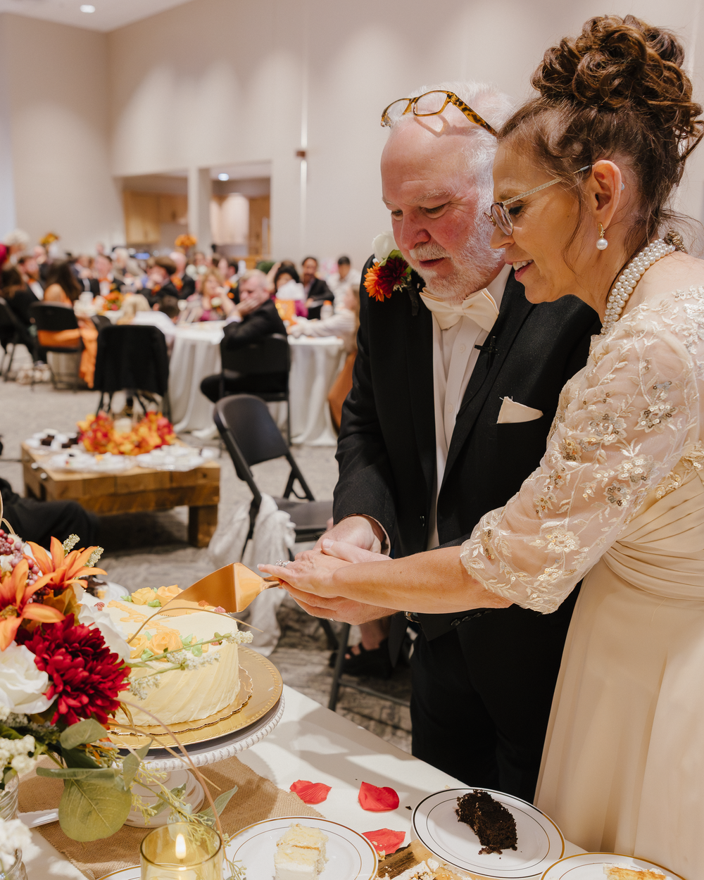 Bride and groom cutting their white wedding cake together at reception with guests seated in background.