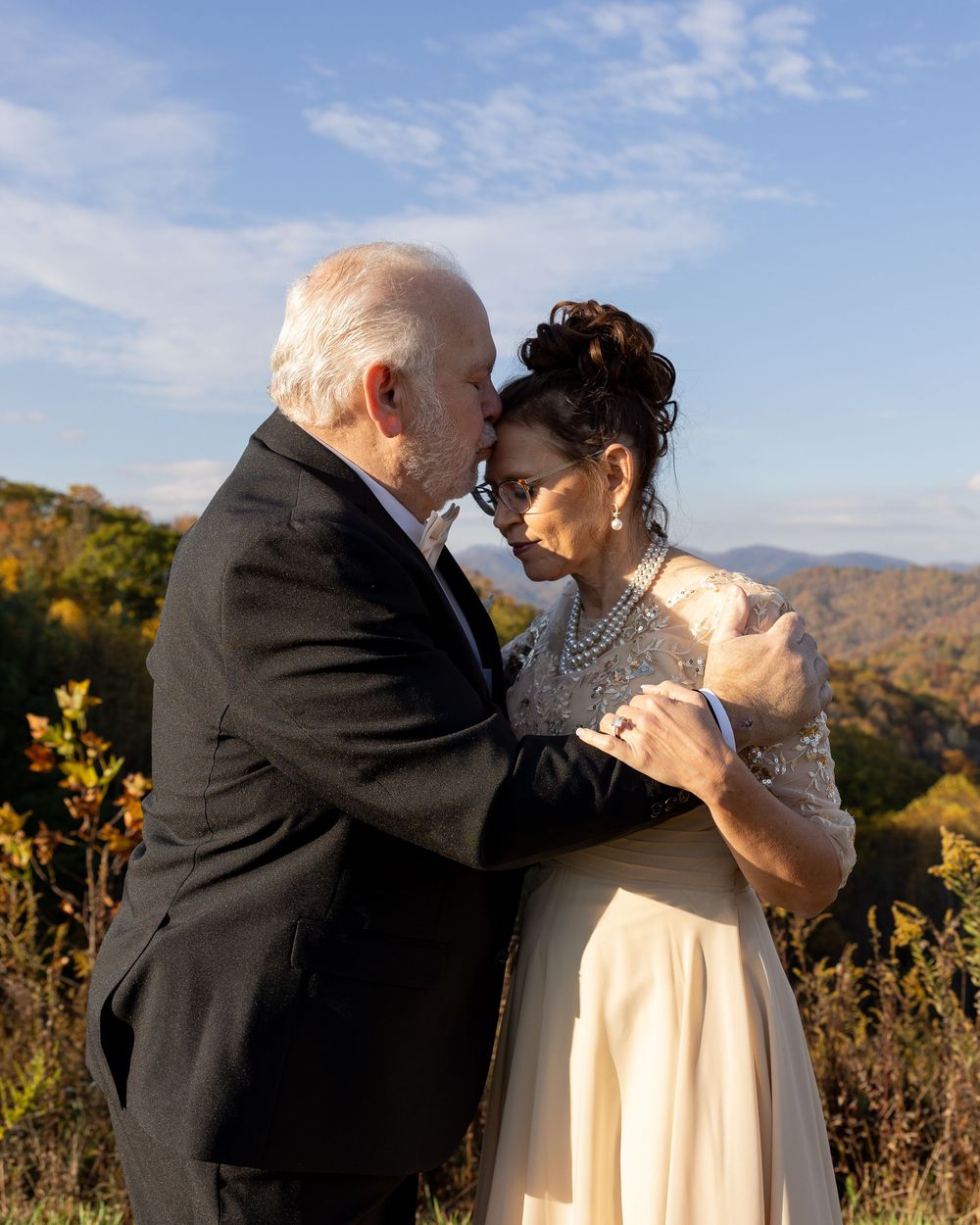 An older couple shares a romantic moment outdoors with colorful autumn mountains in the background.