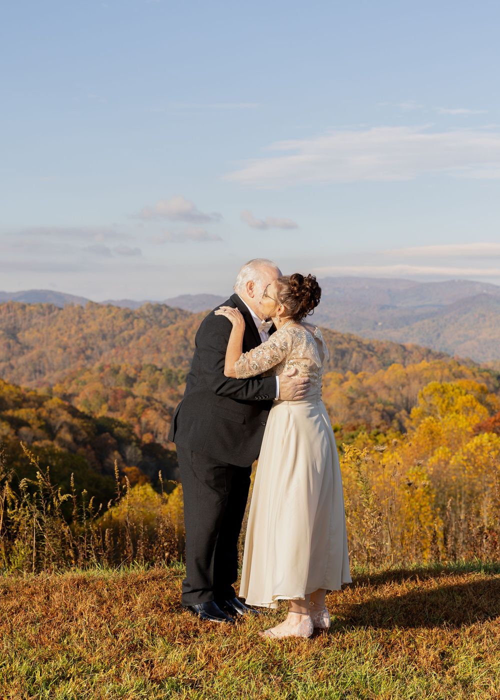 An elderly couple shares a kiss on a hilltop surrounded by vibrant fall foliage and blue sky.