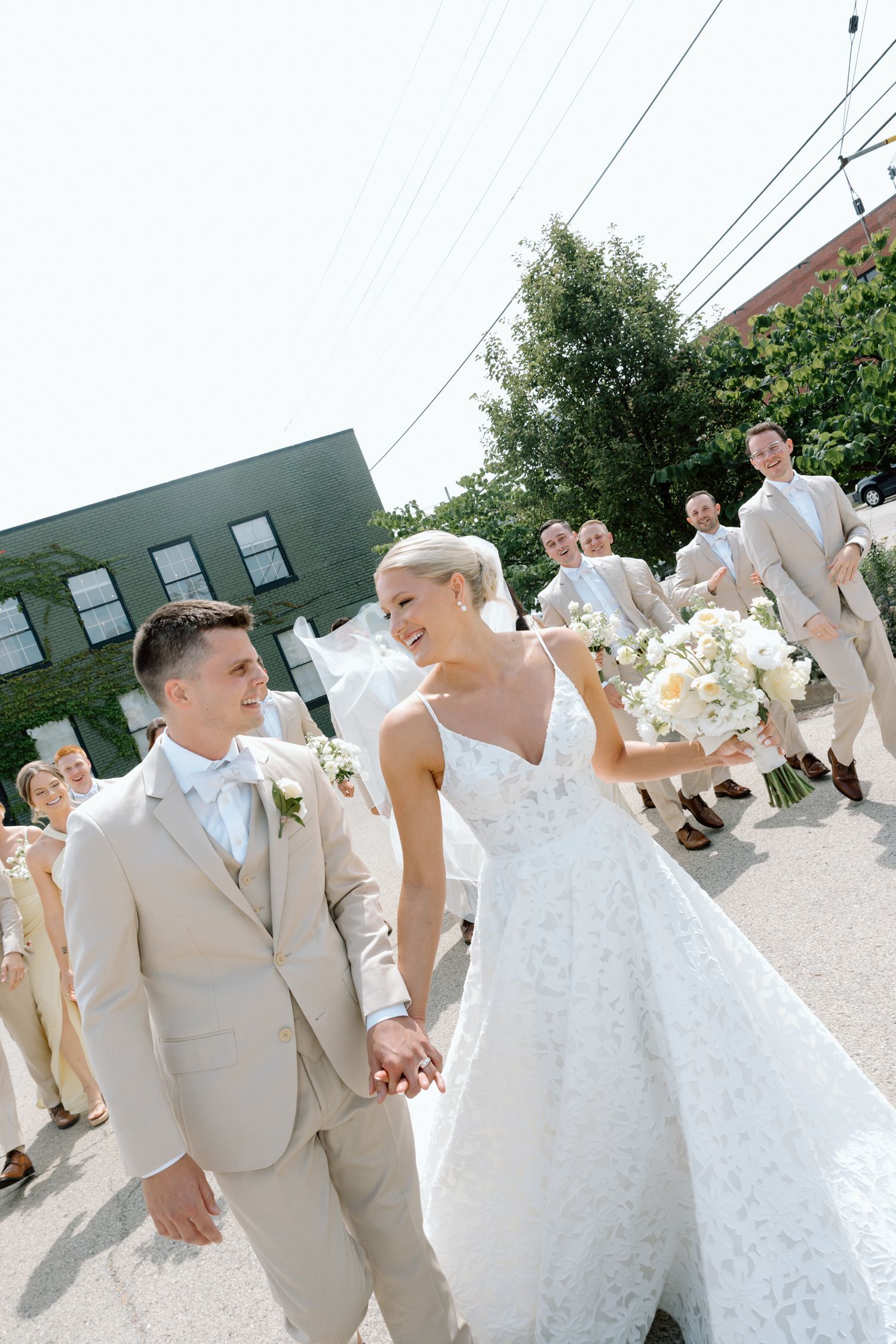 A wedding party in light beige suits and a white dress dancing and celebrating outdoors on a sunny day.