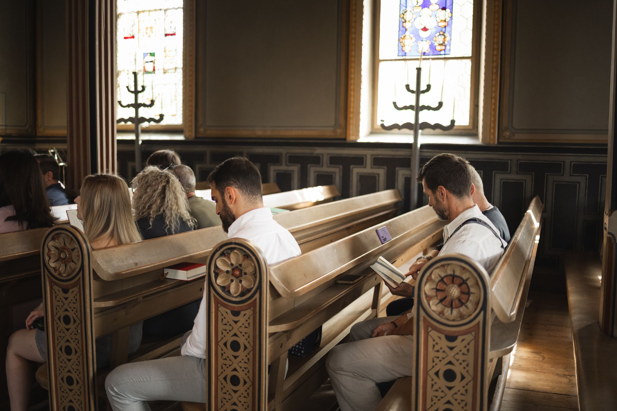 People sit in ornate wooden church pews with decorative carvings and stained glass windows visible in the background.