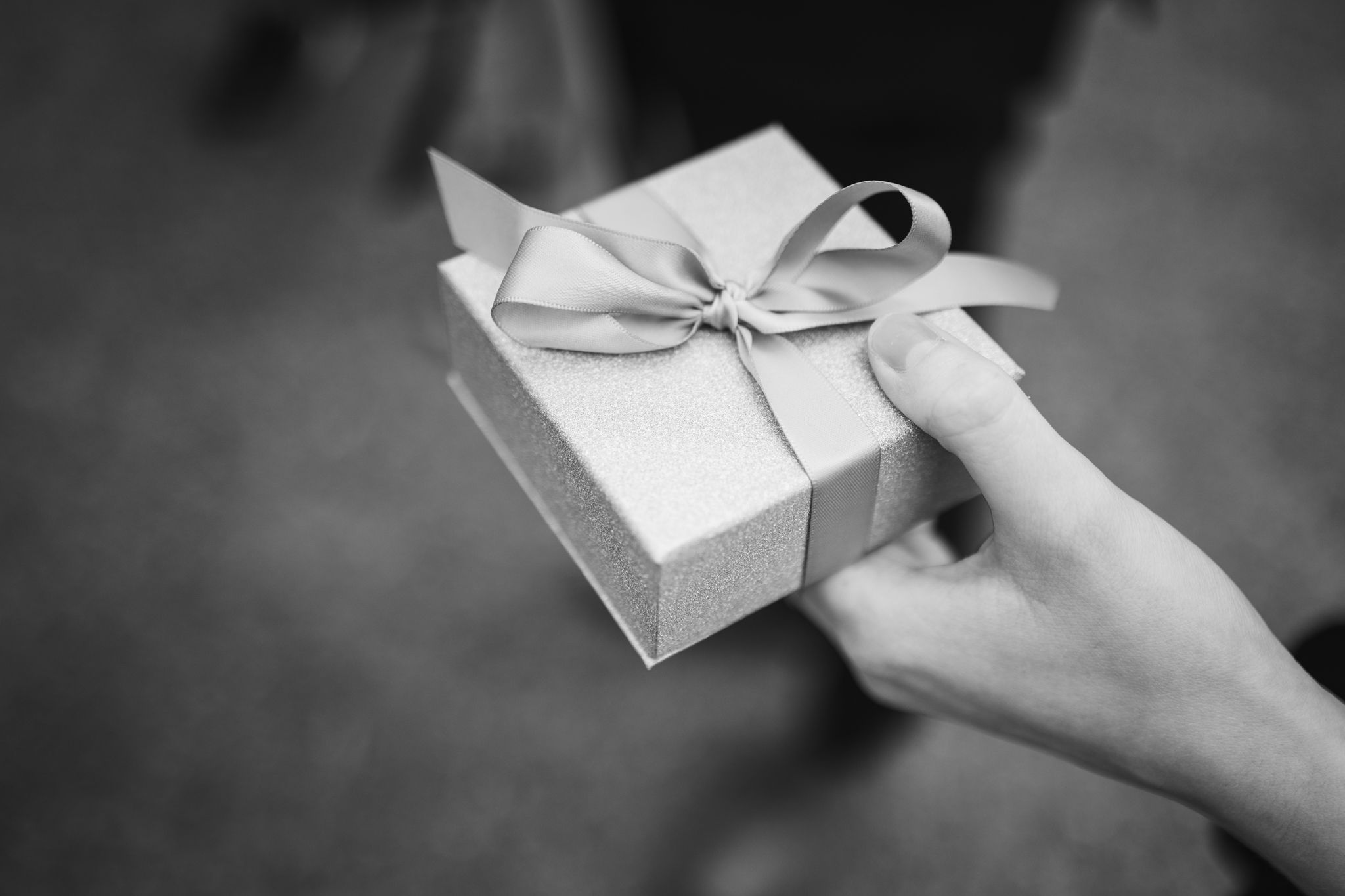 A hand holds an elegant white gift box with a delicate bow ribbon in a black and white photograph.