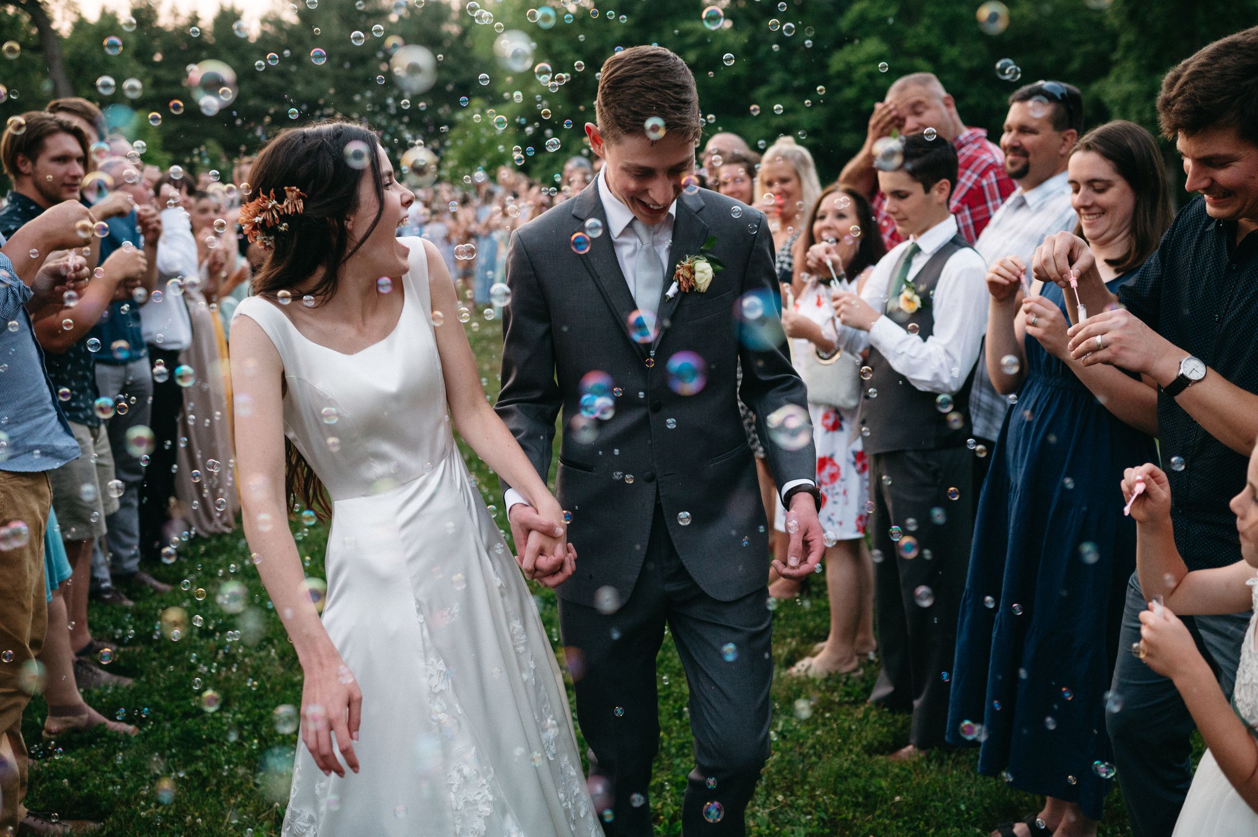 Wedding guests shower newlyweds with bubbles during an outdoor garden celebration at sunset.