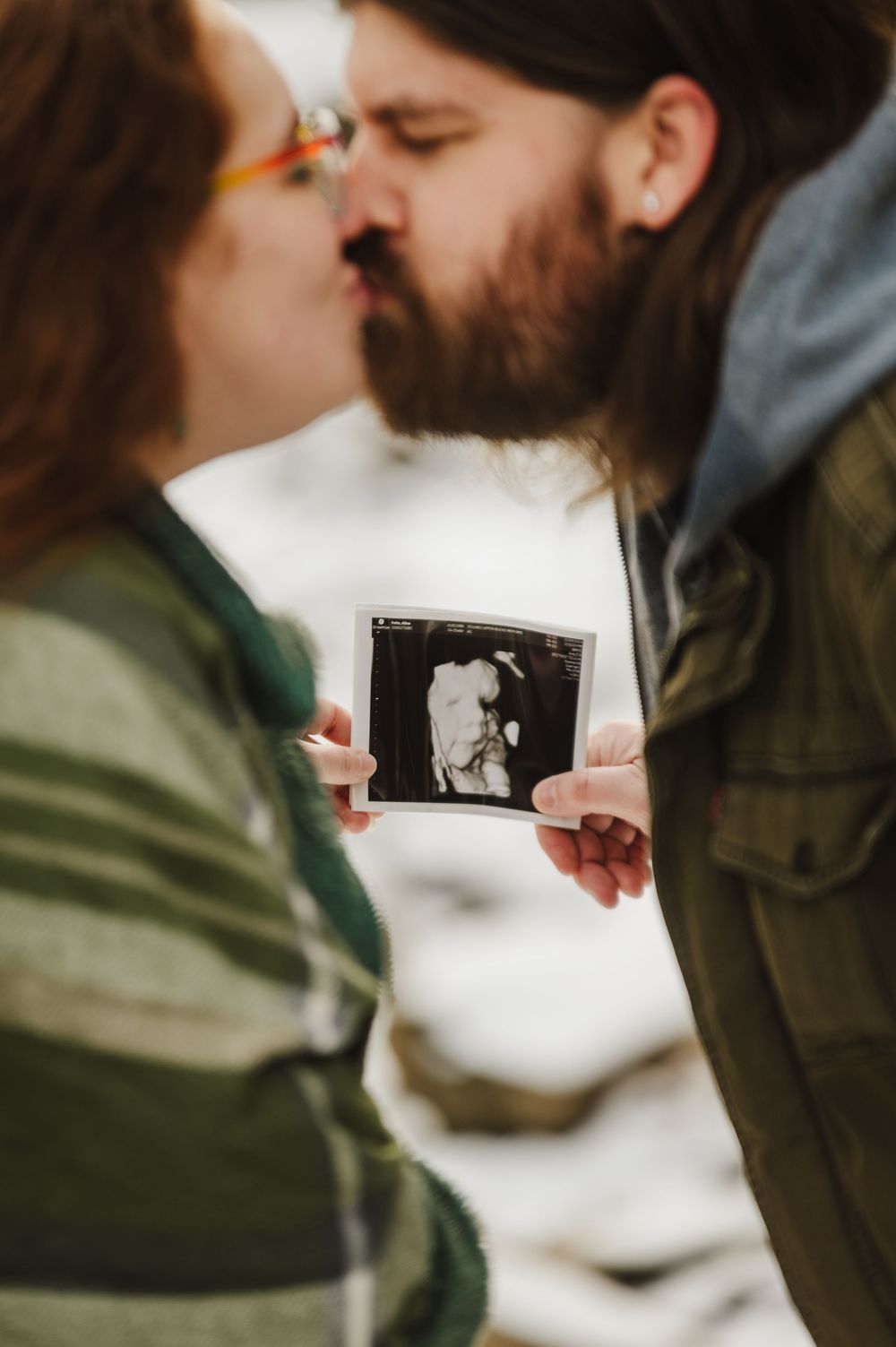 A couple holds up a sonogram photo while sharing an intimate moment outdoors in winter.