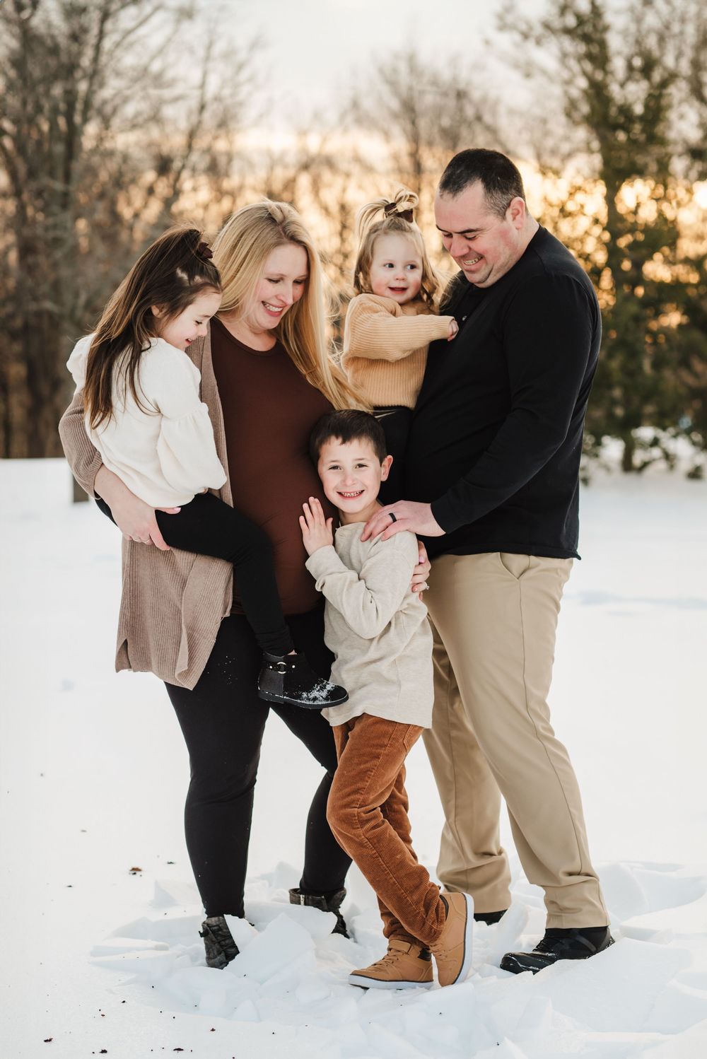 A family of five shares a joyful moment together in the snow during sunset with trees in the background.