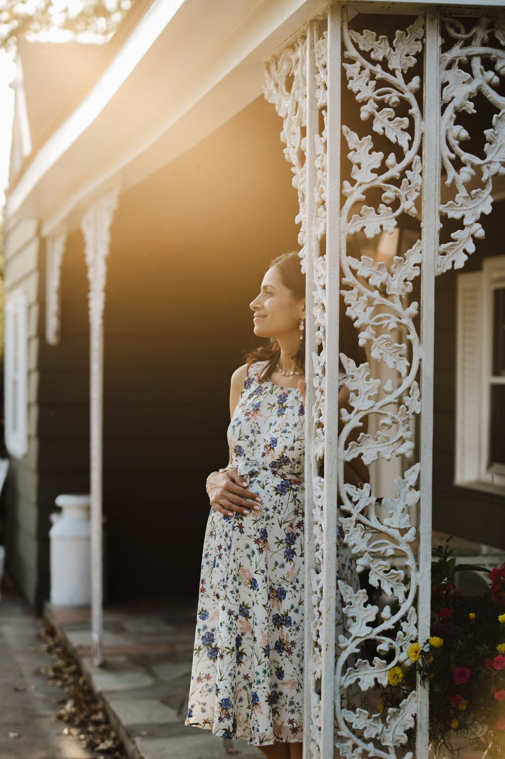A person in a floral dress stands by ornate white Victorian porch pillars at sunset.