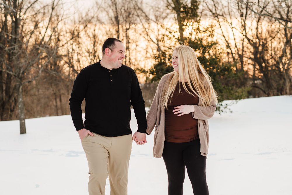 A couple holds hands while walking through a snowy winter landscape at sunset.