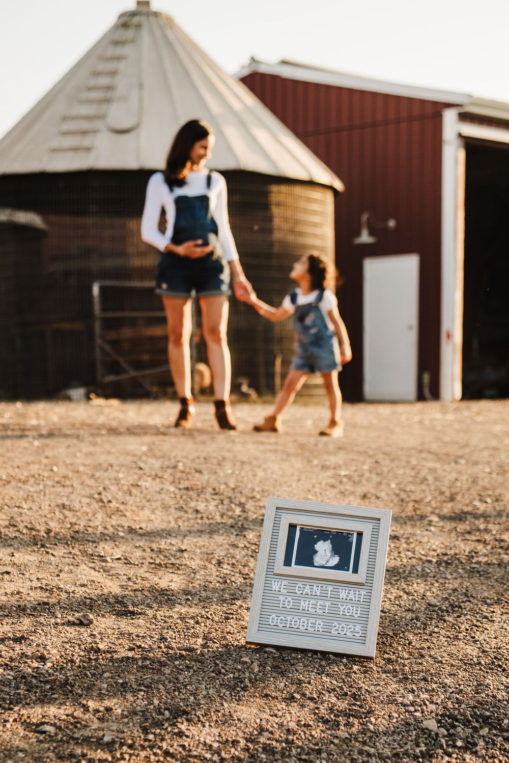Pregnancy announcement photos showing a sign with October 2023 date while figures walk in background near barn buildings.