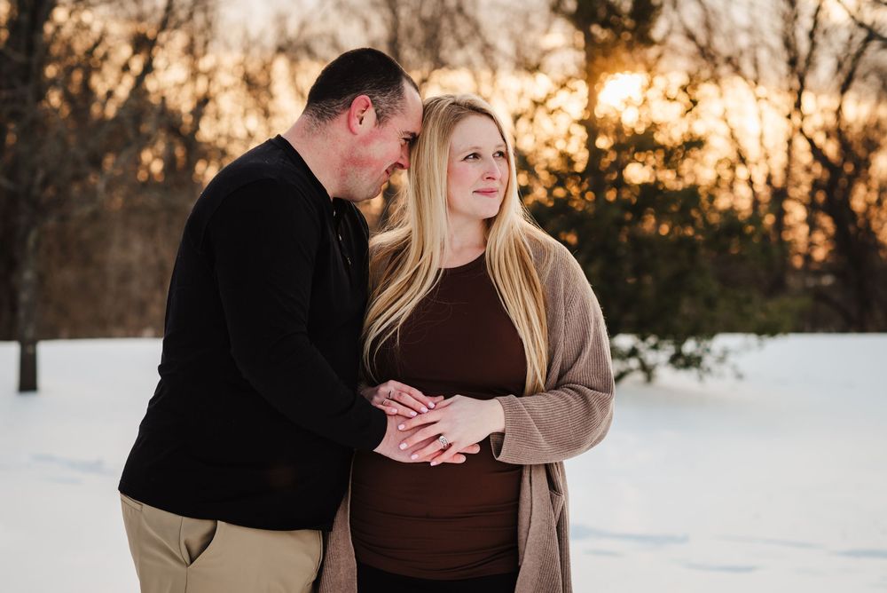 A couple embraces in a snowy winter landscape at sunset with warm golden light filtering through bare trees.