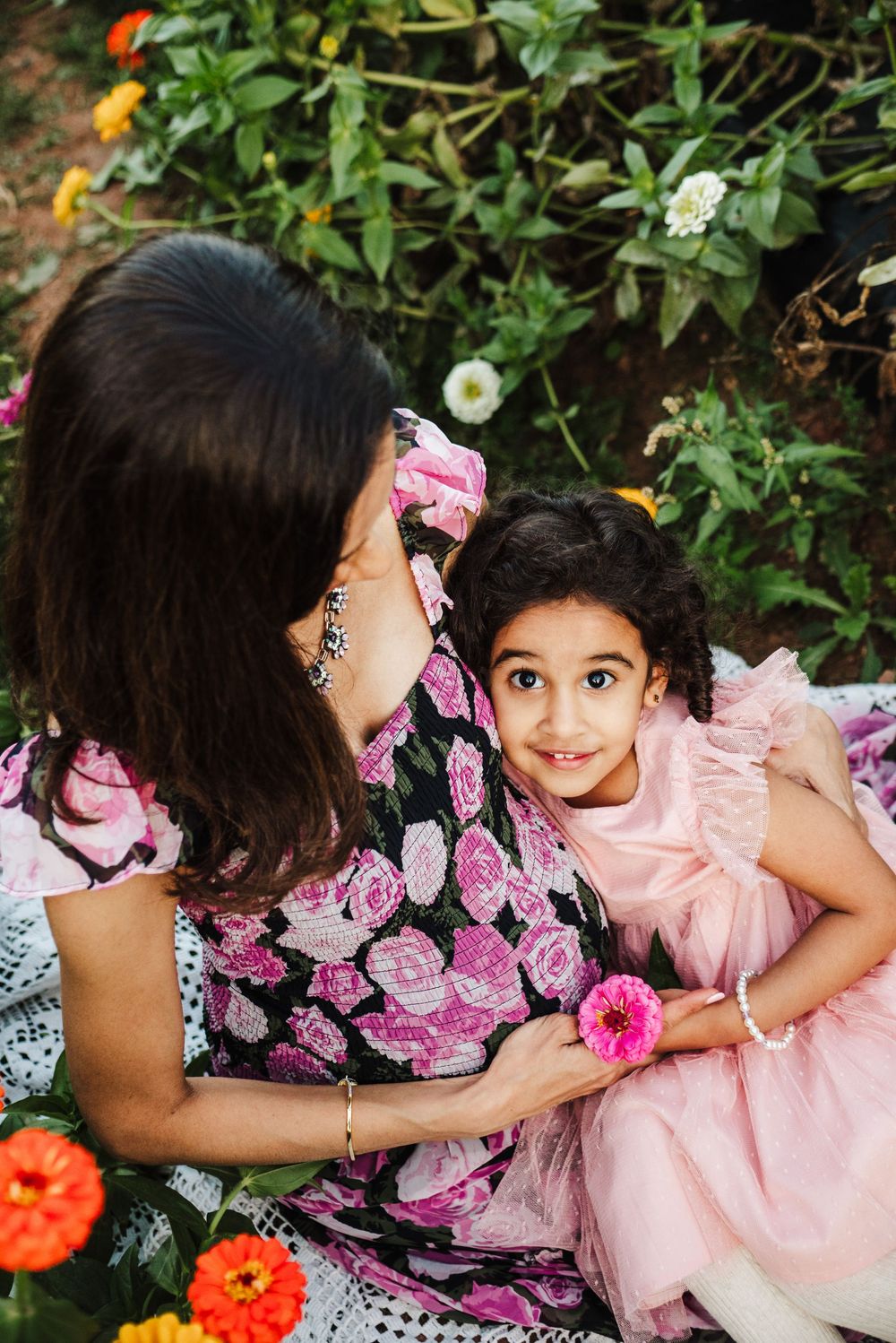 Two people in floral and pink outfits share tender moments in a garden setting with colorful flowers and greenery.