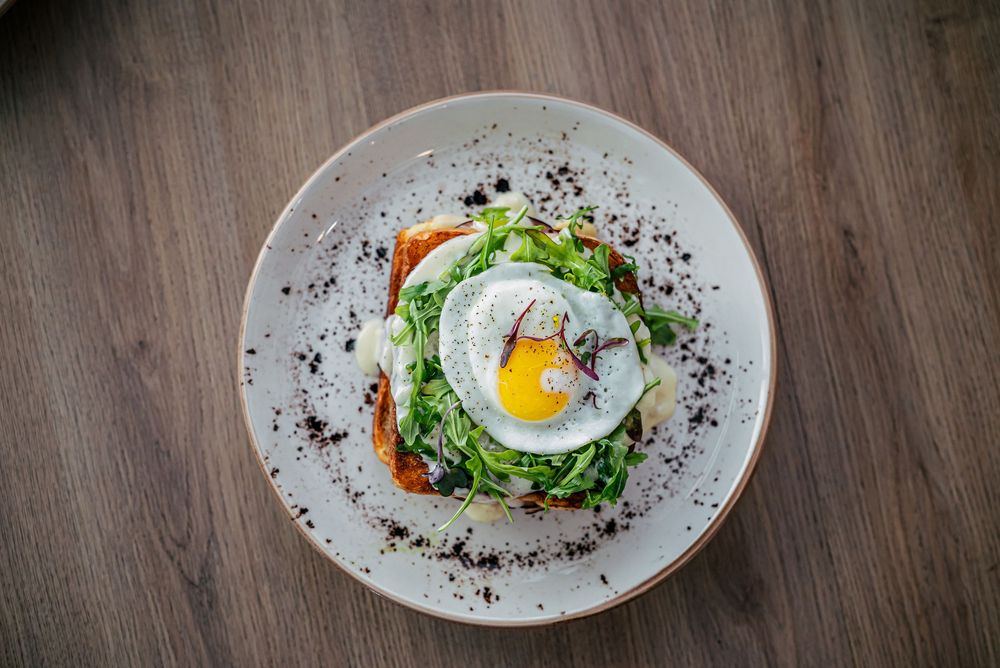 A rustic wooden table displays a series of white plates with sunny side up eggs, fresh greens, and dark seasonings.