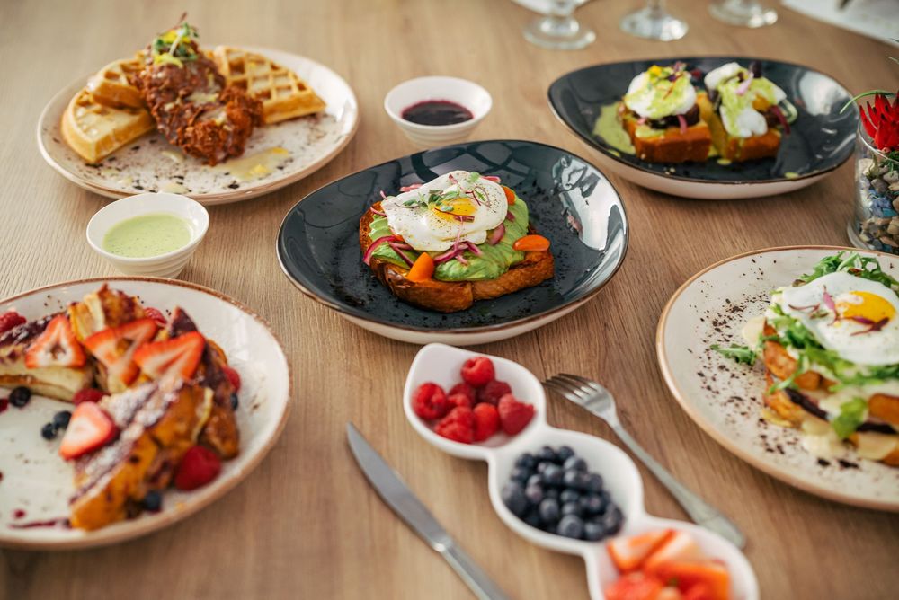 A wooden table set with various brunch dishes including eggs, avocado toast, berries, and shrimp arranged in a row.