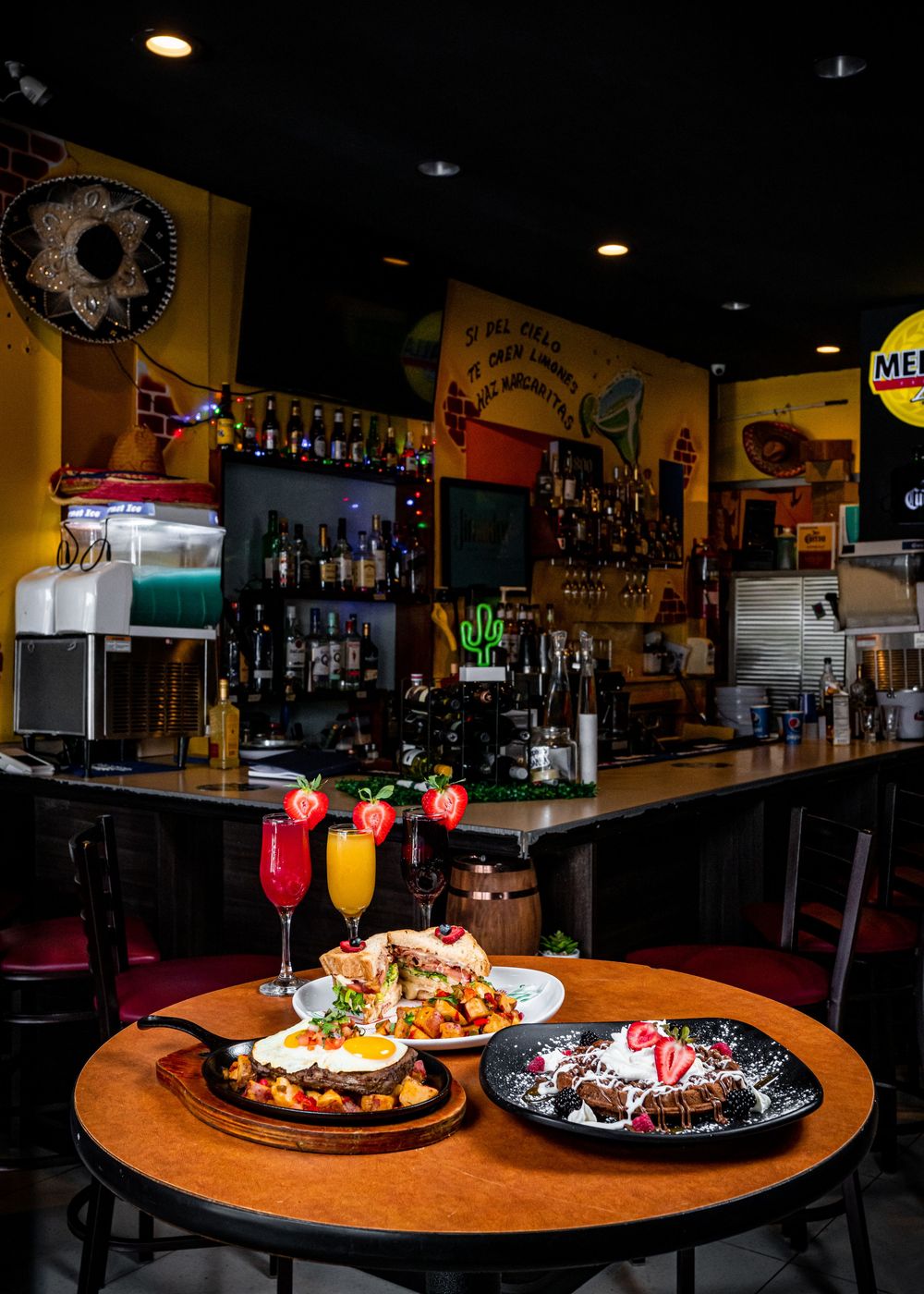 Colorful cocktails and brunch plates served on a wooden table at a bar with bottles and yellow walls in the background.