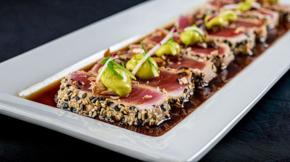 Seared sesame-crusted tuna slices with pepperoncini garnish arranged on a white rectangular plate.
