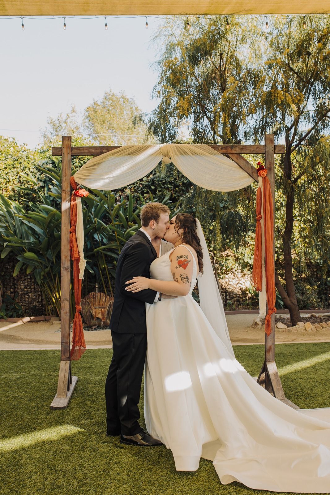 A romantic outdoor wedding ceremony features a wooden arch draped with white fabric on a grassy lawn with trees at River Garden Weddings an.