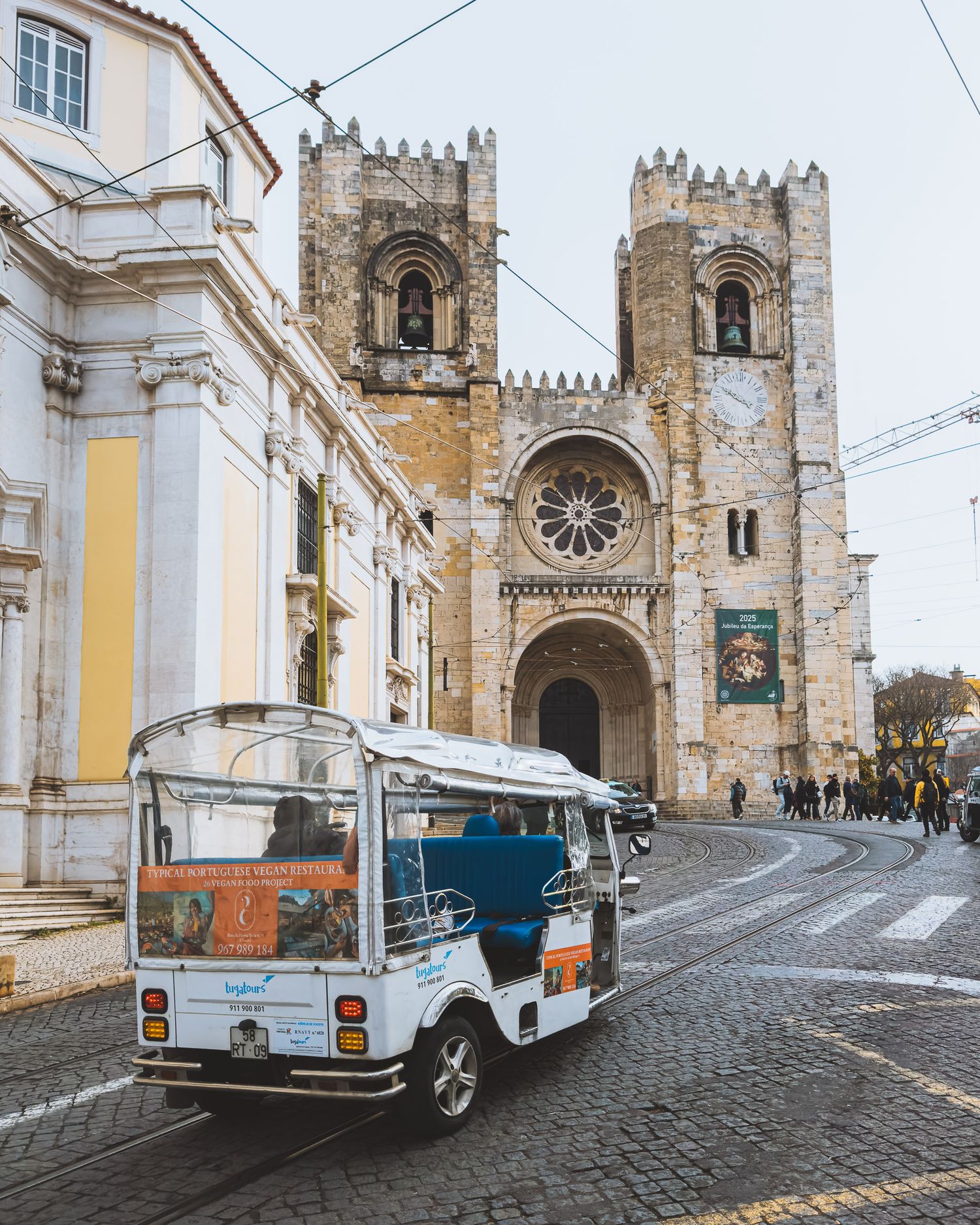 White tourist tuk-tuk vehicles parked in front of a grand Gothic cathedral with twin towers. Lisbon.