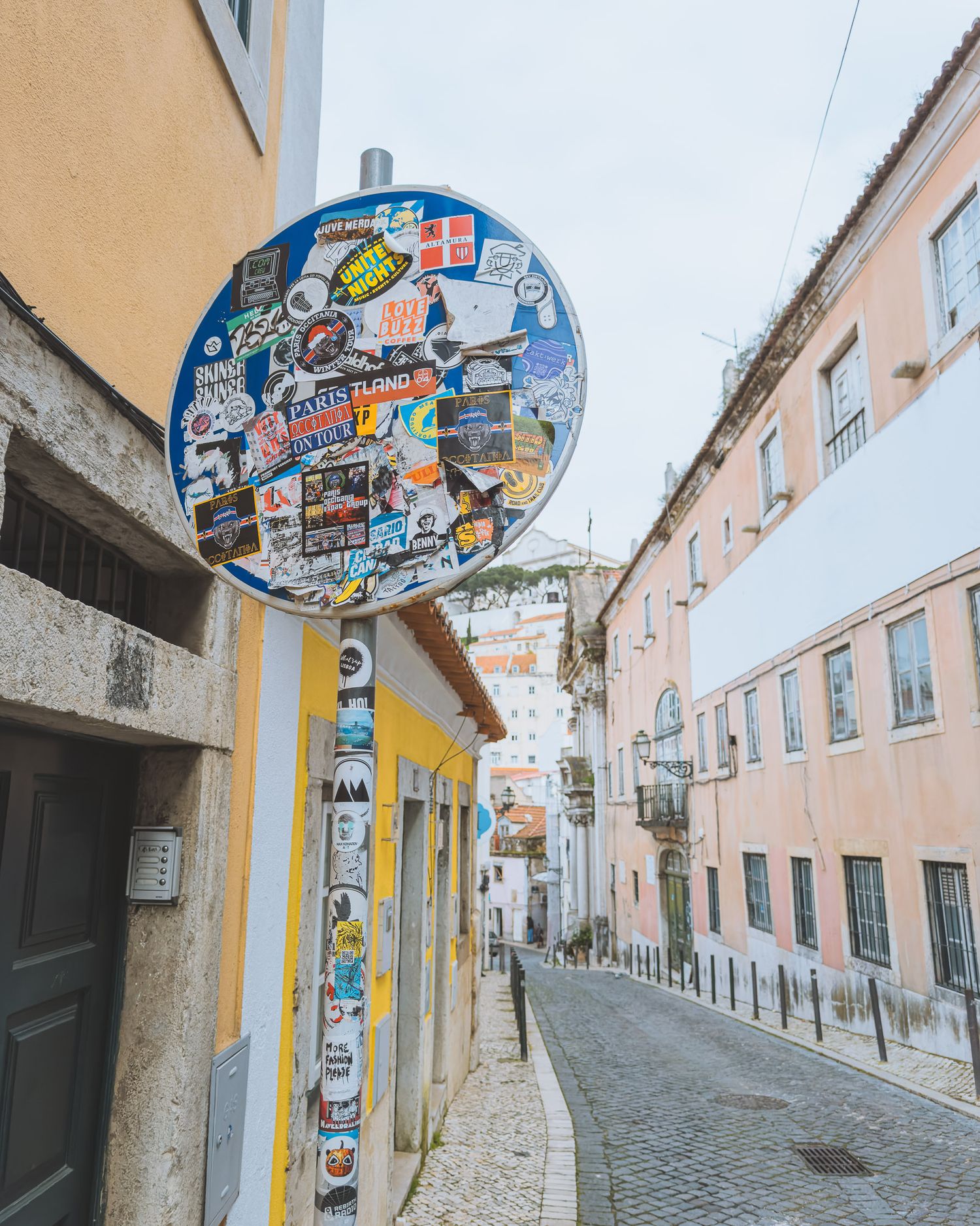 Sticker-covered street sign on yellow wall along narrow cobblestone street in old European city.