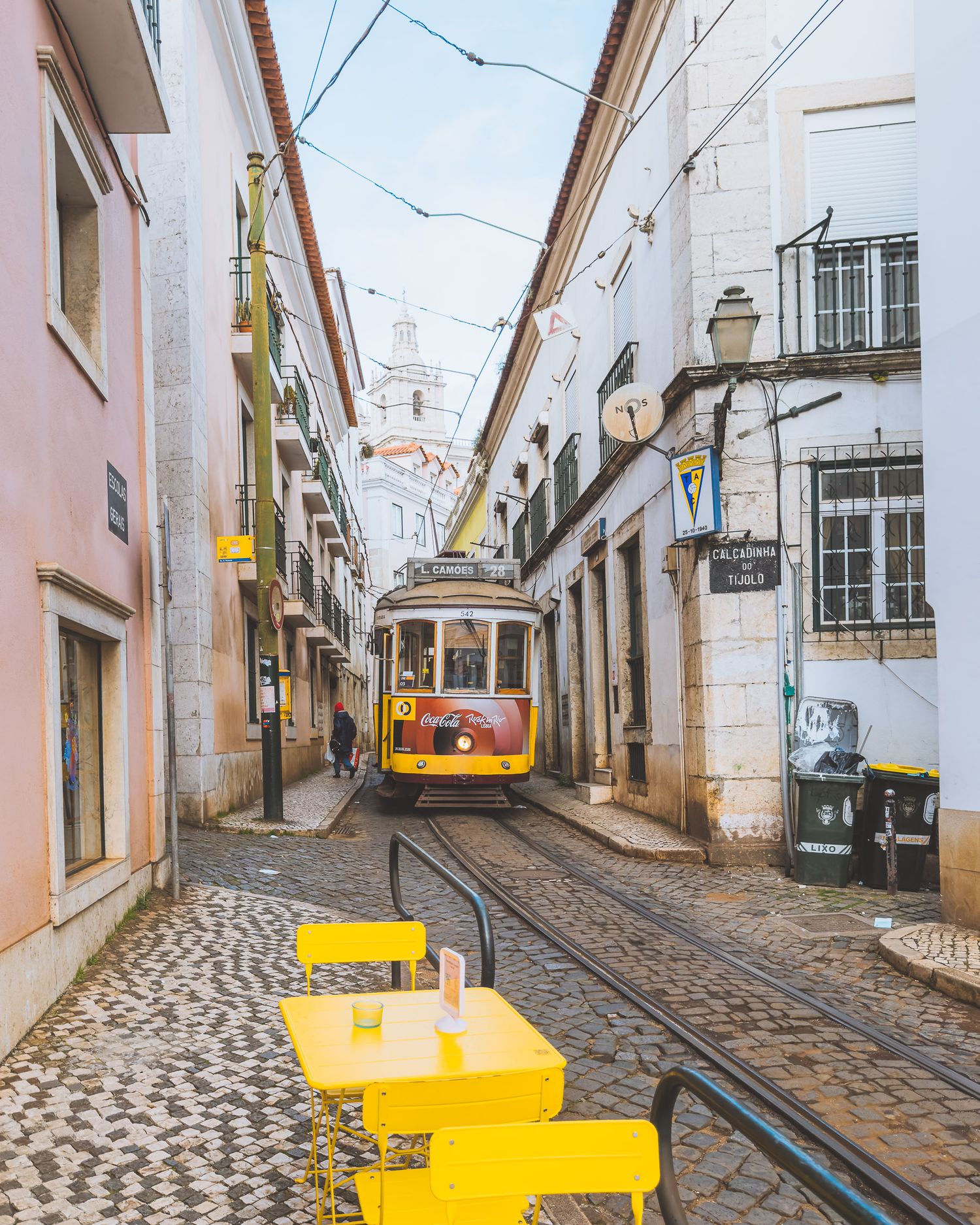Yellow vintage tram on cobblestone street with outdoor cafe tables in Lisbon.