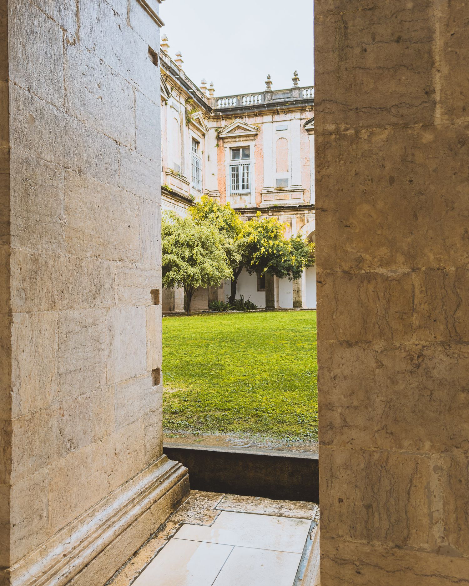 Stone colonnade walkway leading to a green courtyard with classical architecture.