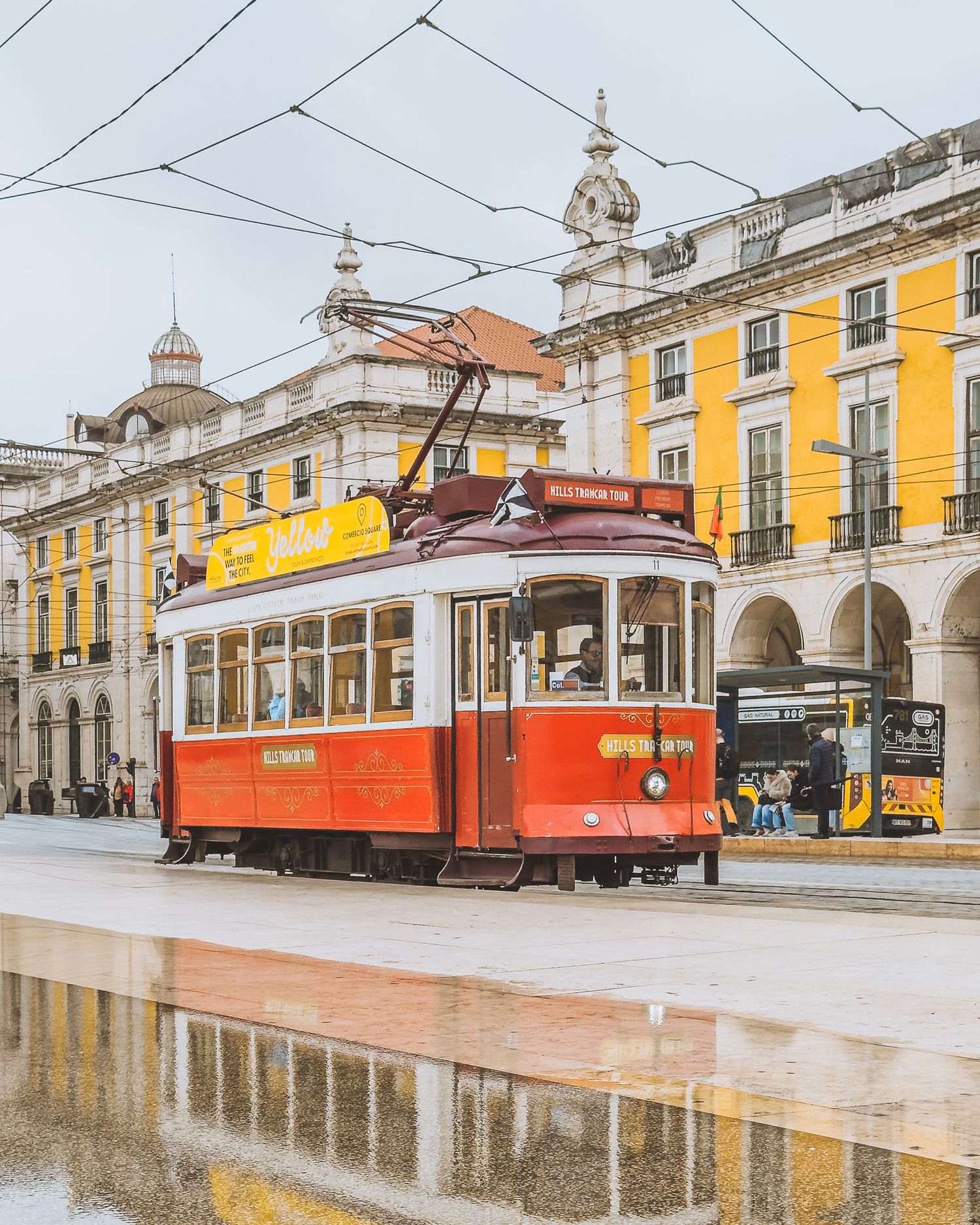 Historic red and yellow trams parked at a station with classical architecture and wet pavement in Lisbon.