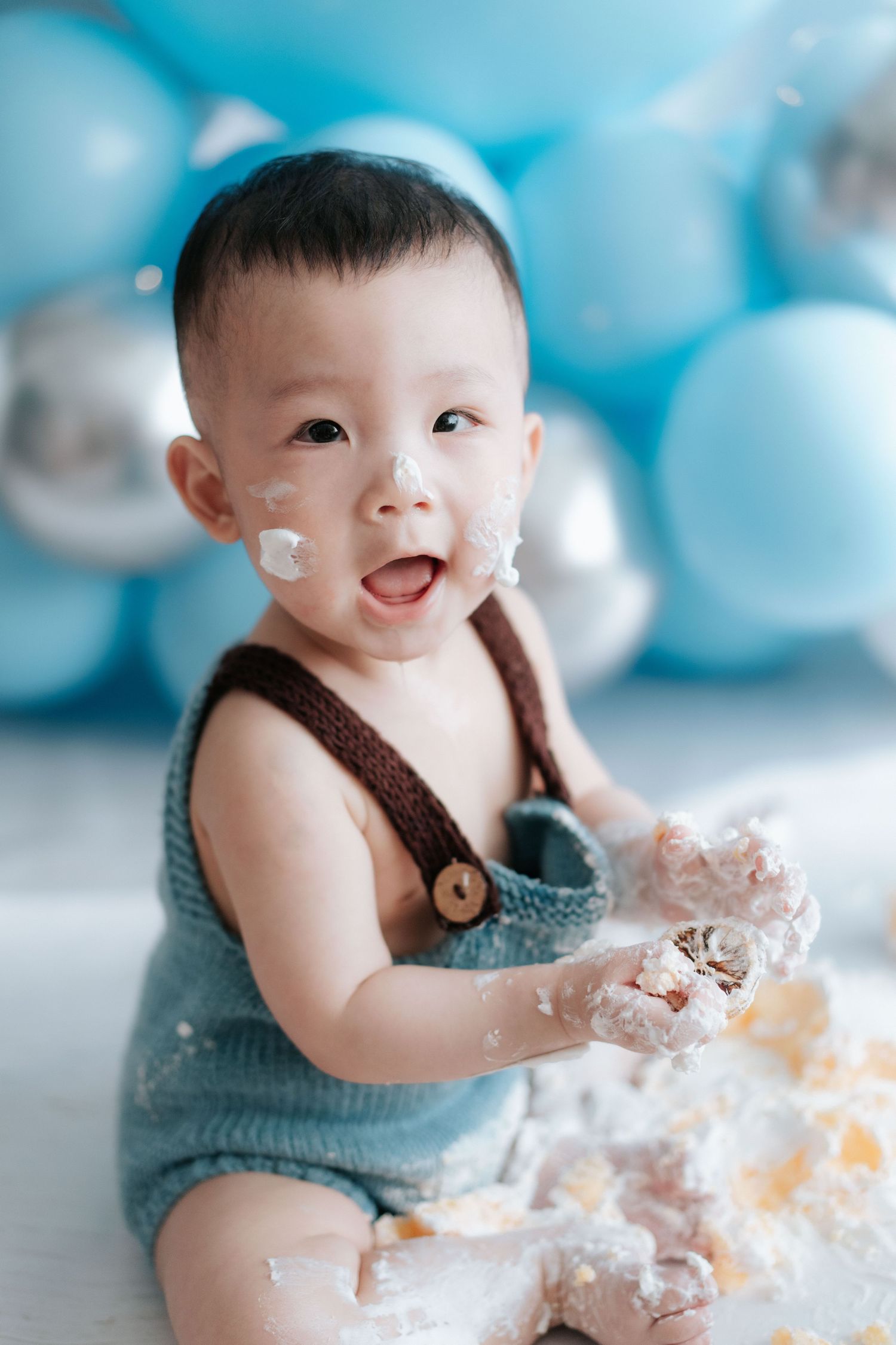 A baby wearing a blue knit outfit smiles and plays with cake during a cake smash photo session.