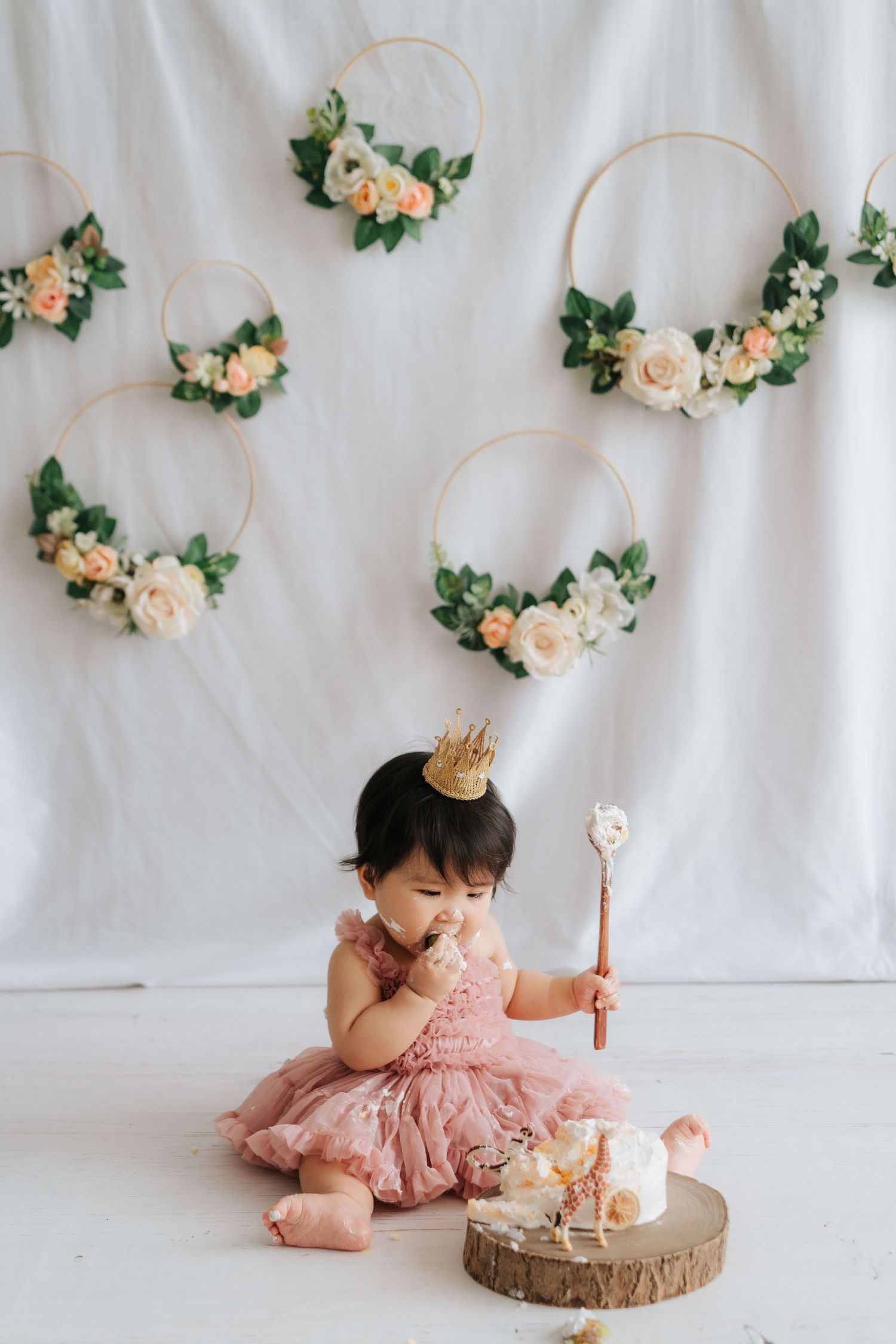 A baby in a pink tutu dress sits on a white backdrop with floral decorations while having a cake smash photo session.
