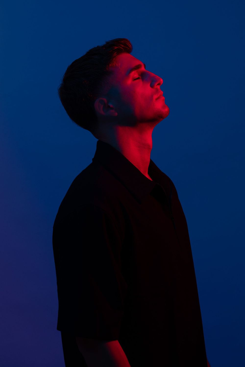 A young man in a black shirt poses against a blue background, illuminated by dramatic red lighting.