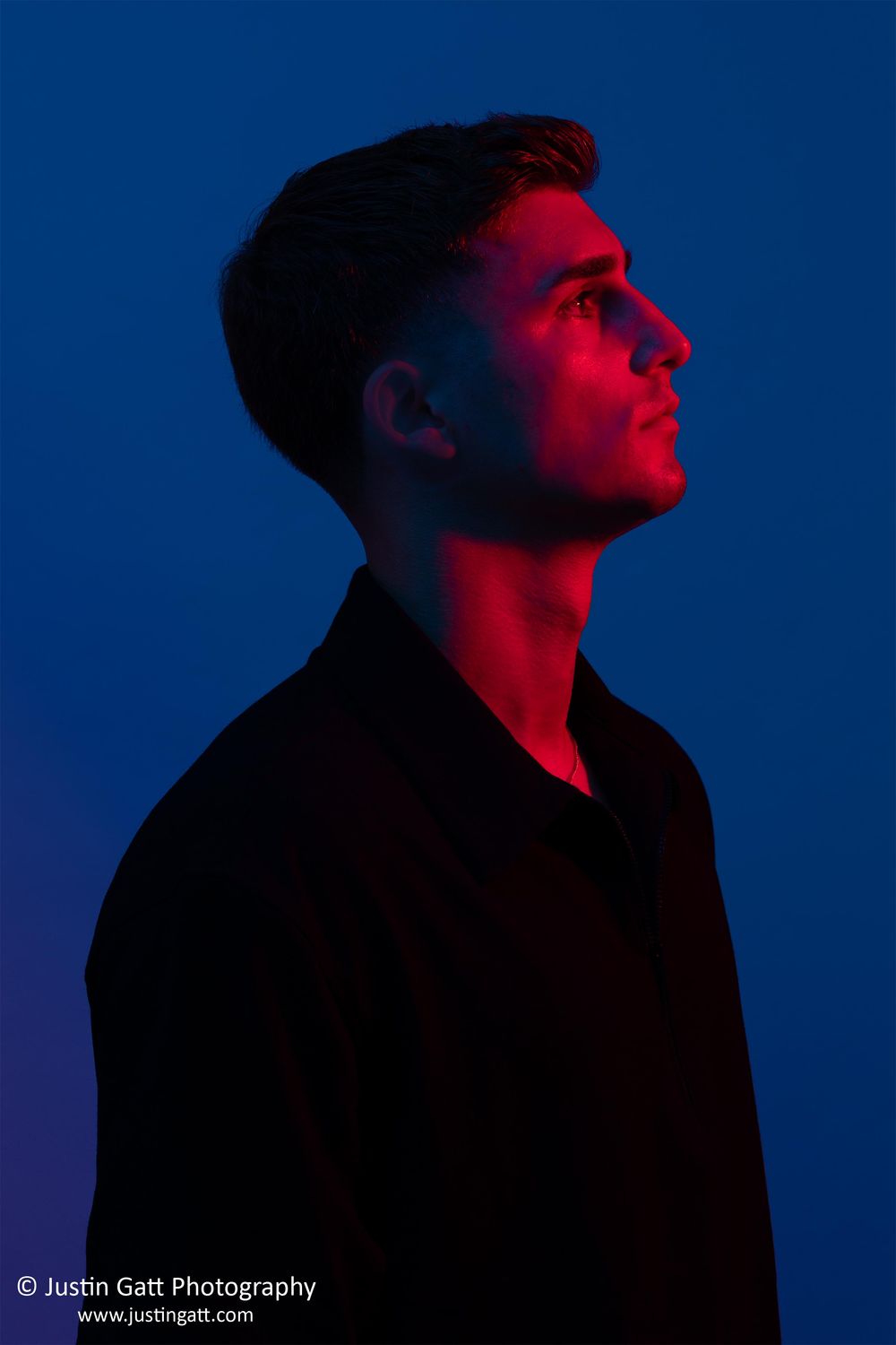 Young man in profile view bathed in dramatic red and blue lighting against a deep blue background.