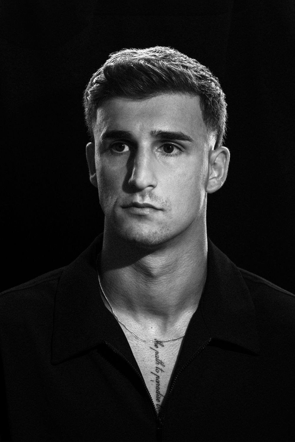 Black and white portrait of a young man with short hair, wearing an open collar shirt with a chest tattoo.