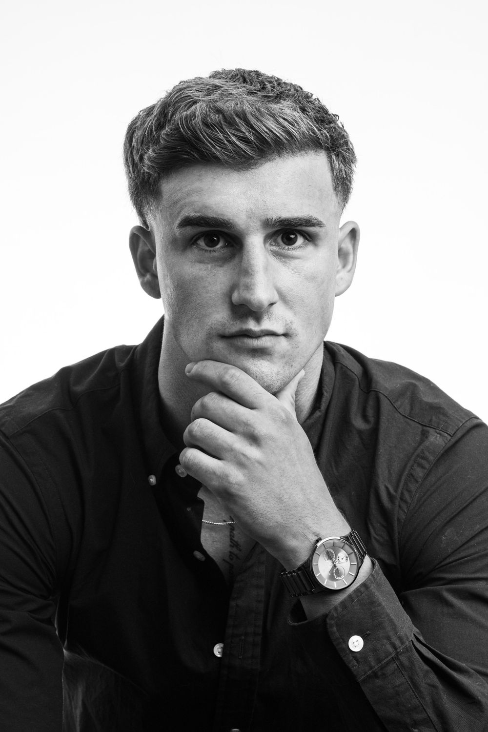 Black and white portrait of a young man in a dark shirt, hand on chin, wearing a watch, thoughtful expression.