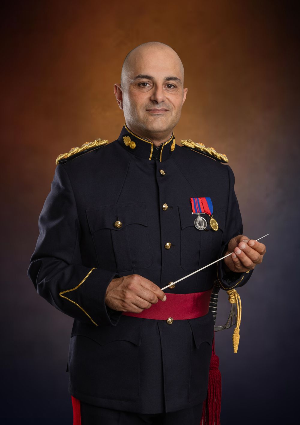 Military conductor in dress uniform holding a baton, wearing medals and gold epaulettes.