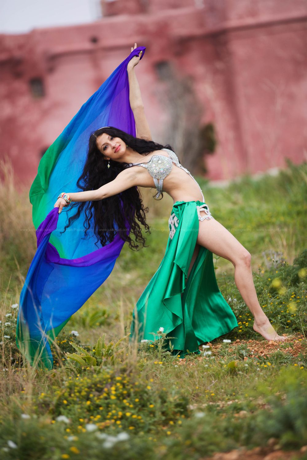 Dancer in green and blue flowing fabrics poses gracefully in a meadow near a pink wall.